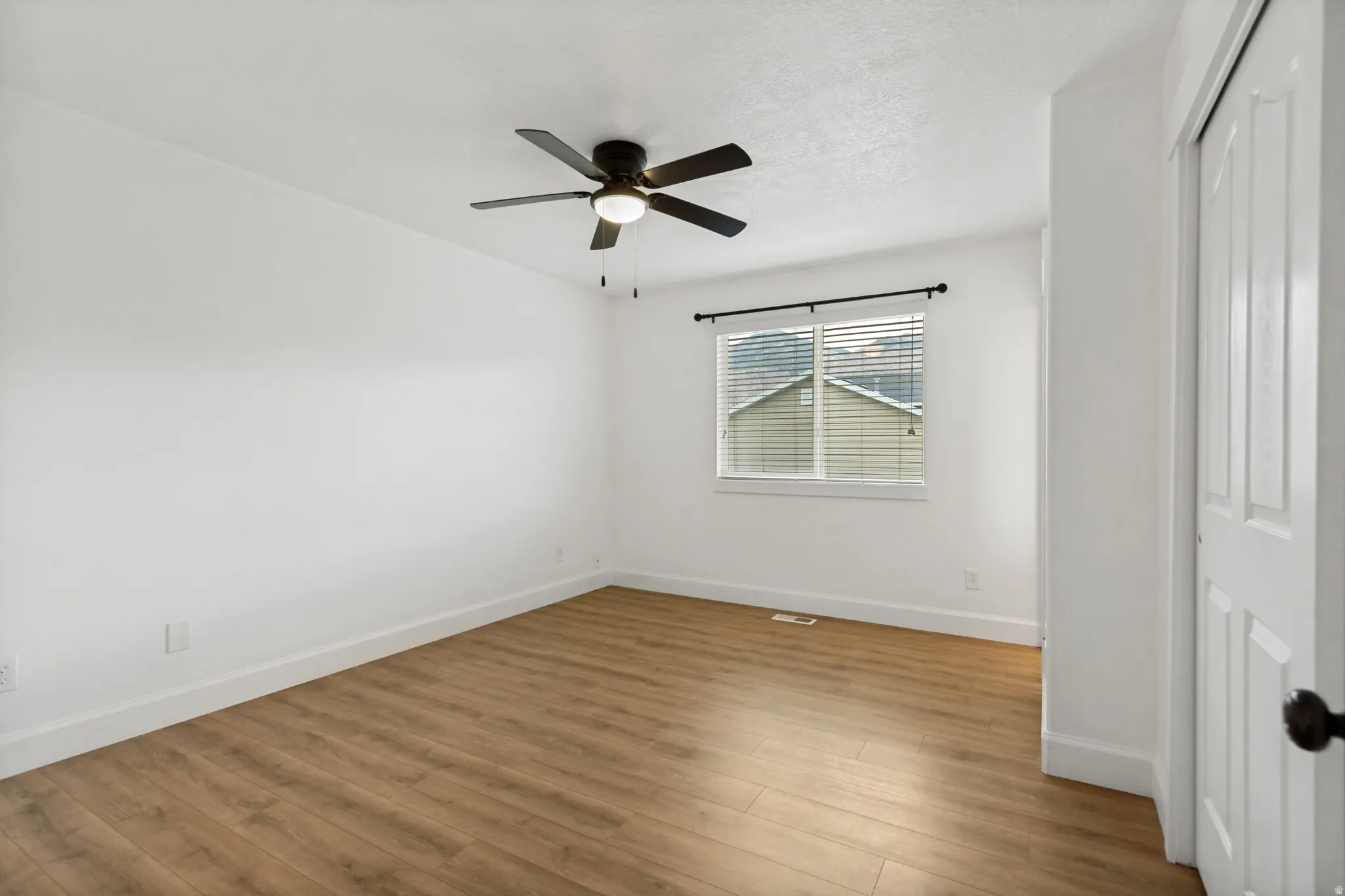 Spacious room featuring wood-finish flooring, a ceiling fan with integrated lighting, window with blinds, and a paneled closet door