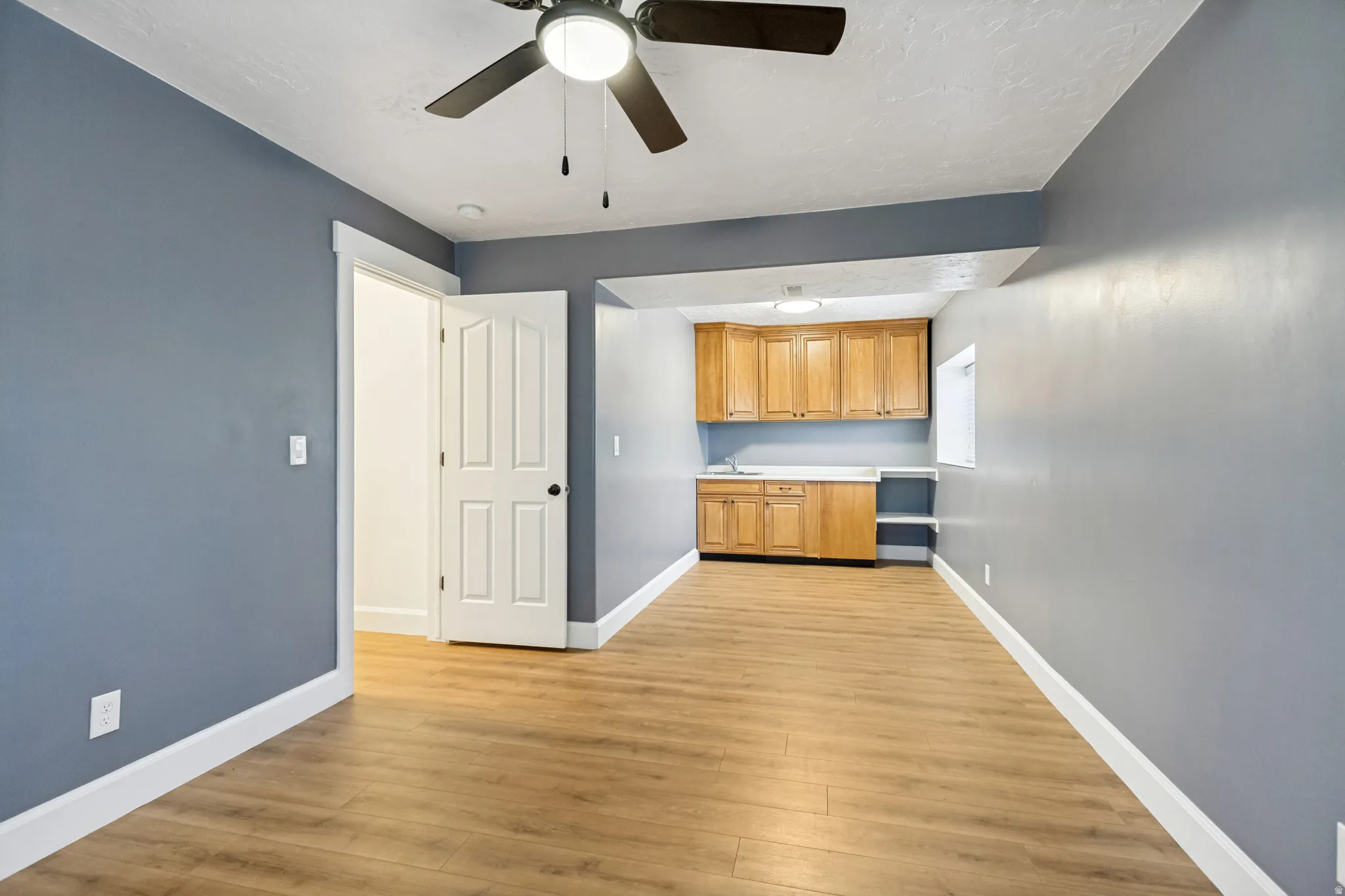 Wood-finish flooring, ceiling fan, white paneled doors, light wood cabinetry, and a compact sink area
