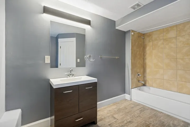 Bathroom featuring a dark wood-finish vanity with an integrated white sink, a vanity mirror with overhead lighting, a bathtub with tiled surround, and wood-finish flooring