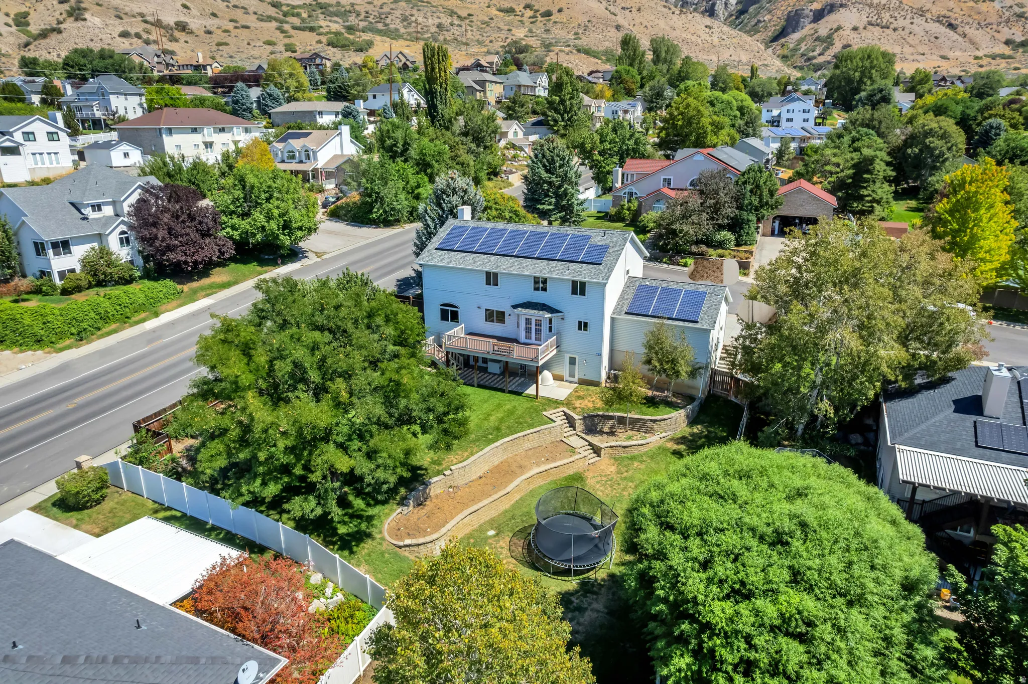 Two-story residence with multiple solar panel installations, a ground-level deck, and a terraced backyard featuring a sloped retention wall