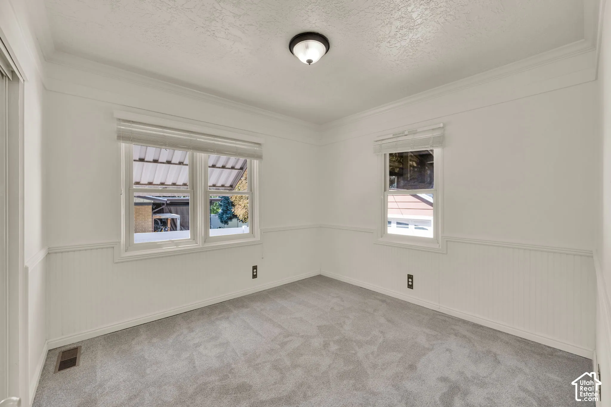 Light gray carpeting, crown molding, two windows with blinds, and white wainscoting
