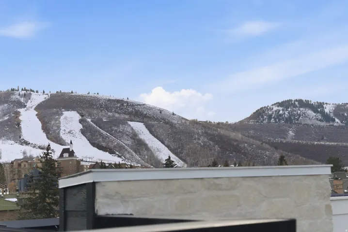 Snow-covered mountain landscape with visible ski runs, surrounded by evergreen trees and distant buildings