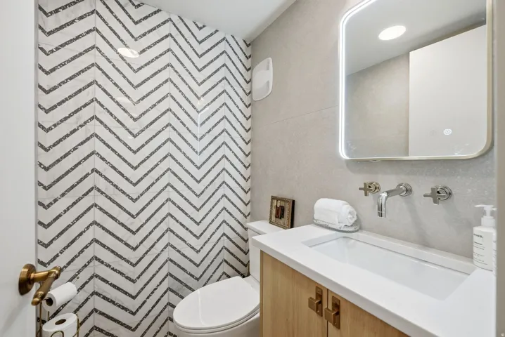 Herringbone tile accent wall, integrated vanity sink, wood-finish cabinetry with brass hardware, wall-mounted faucet, and an illuminated mirror