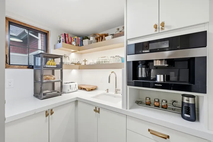 Built-in Miele coffee system, undermount sink with a gooseneck faucet, white cabinetry with brass hardware, wood-framed window, and integrated shelving with under-shelf lighting