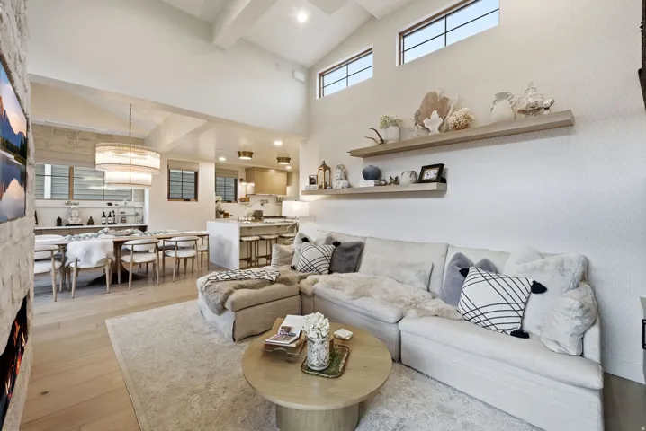 Vaulted ceilings with exposed beams, high transom windows, a stone-clad fireplace, wood-finish flooring, and floating wall shelves