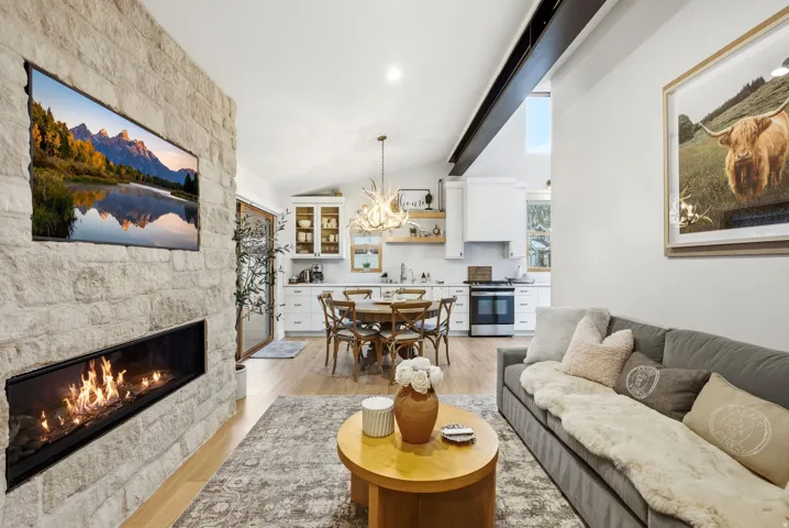 Stone-clad fireplace with a linear firebox, vaulted ceiling with exposed beam, wood-finish flooring, white kitchen cabinetry, and built-in stainless steel appliances