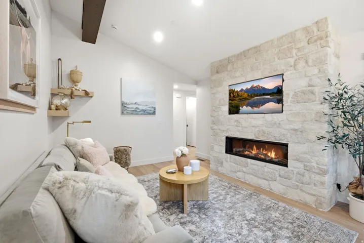 Vaulted ceiling with exposed wood beam, stone accent wall with integrated linear fireplace, recessed lighting, and wood-finish flooring