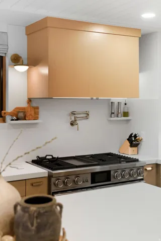 Stainless steel range with griddle, wood-finish range hood, wall-mounted pot filler, white countertops, and light wood-finish cabinetry