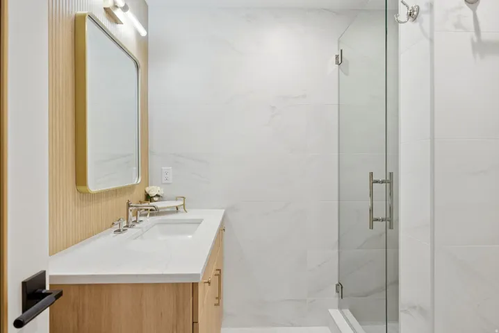 Bathroom featuring a wood-finish vanity with an integrated sink, a gold-framed mirror, polished chrome fixtures, a glass-enclosed shower, and large format wall tiling