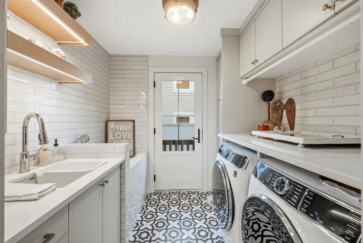 Utility room with patterned floor tile, white subway tile backsplash, light wood open shelving with integrated lighting, a stainless steel sink with a gooseneck faucet, and light-toned cabinetry