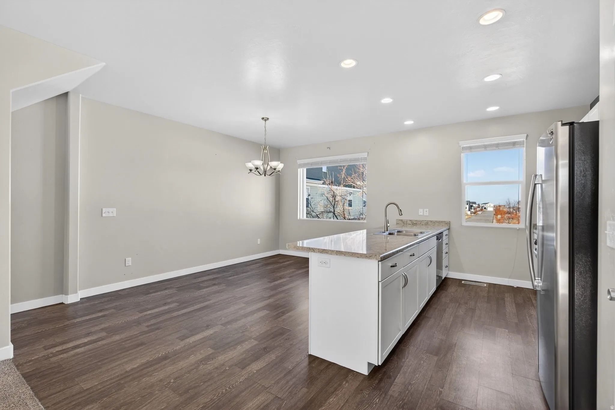 Open-concept living space with wood-finish flooring, recessed lighting, a kitchen island with a sink, stainless steel refrigerator, and a dining area with a chandelier