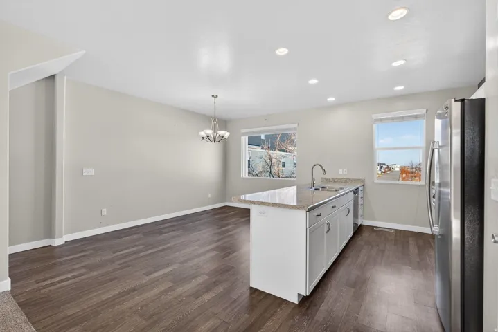 Open-concept living space with wood-finish flooring, recessed lighting, a kitchen island with a sink, stainless steel refrigerator, and a dining area with a chandelier