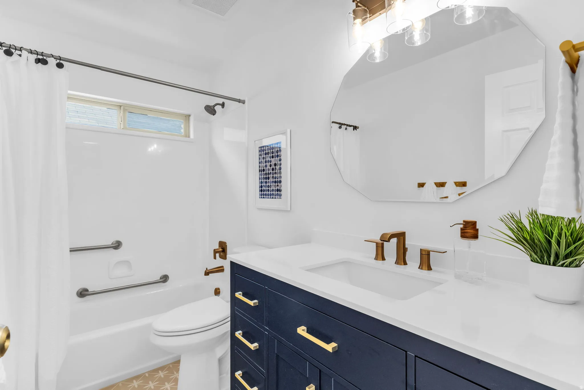 Bathroom featuring a white tub with grab bars, a dual-flush toilet, a navy blue vanity with gold-tone hardware, a white countertop with an undermount sink, and an octagonal mirror