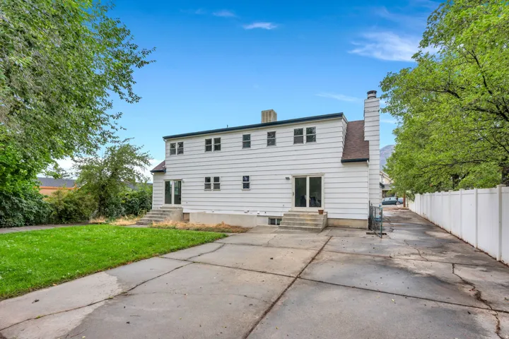 White exterior siding with multiple windows, two ground-level exterior doors with concrete steps, a chimney, and an expansive concrete patio