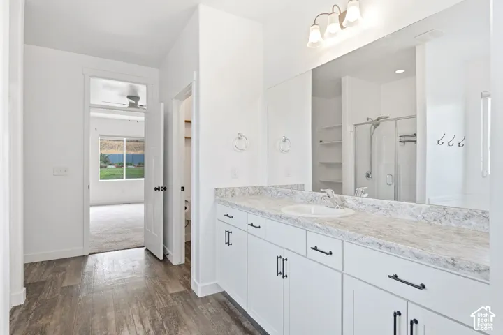 Dual-sink vanity with a light-toned countertop, wood-finish flooring, white cabinetry with dark hardware, a spacious mirror, and a shower enclosure