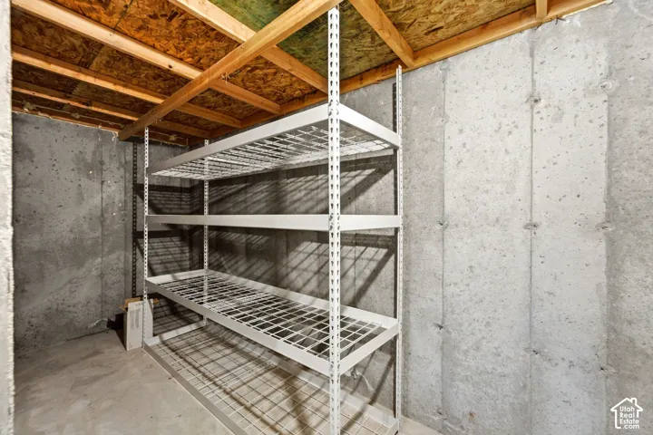 Unfinished basement space featuring concrete walls, concrete flooring, exposed wood ceiling joists, and a built-in metal shelving unit