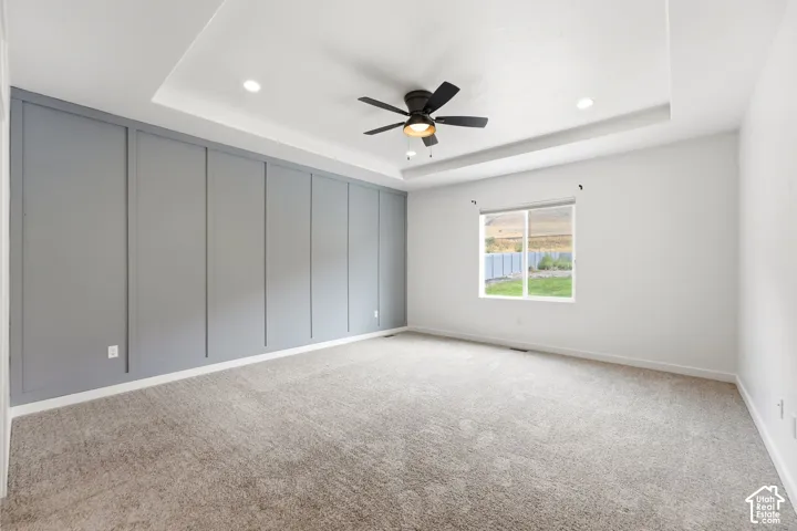 Tray ceiling with recessed lighting and a ceiling fan, accent wall with vertical paneling, neutral carpeting, and a window overlooking a fenced yard
