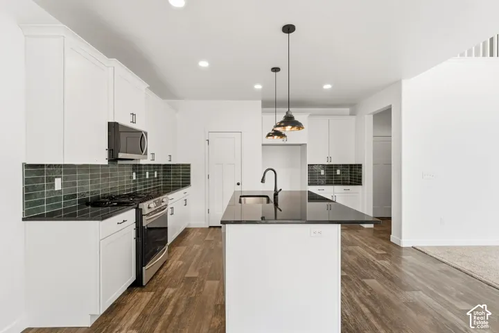 Kitchen featuring a central island with a sink, pendant lighting, stainless steel appliances, white cabinetry, and dark subway tile backsplash