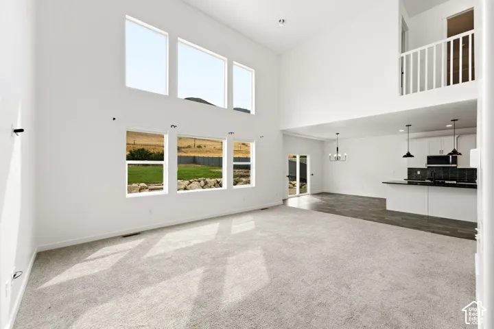 Two-story living area with high ceilings, large windows, carpet flooring, a visible kitchen area with a peninsula, and an upper-level balcony with white railings