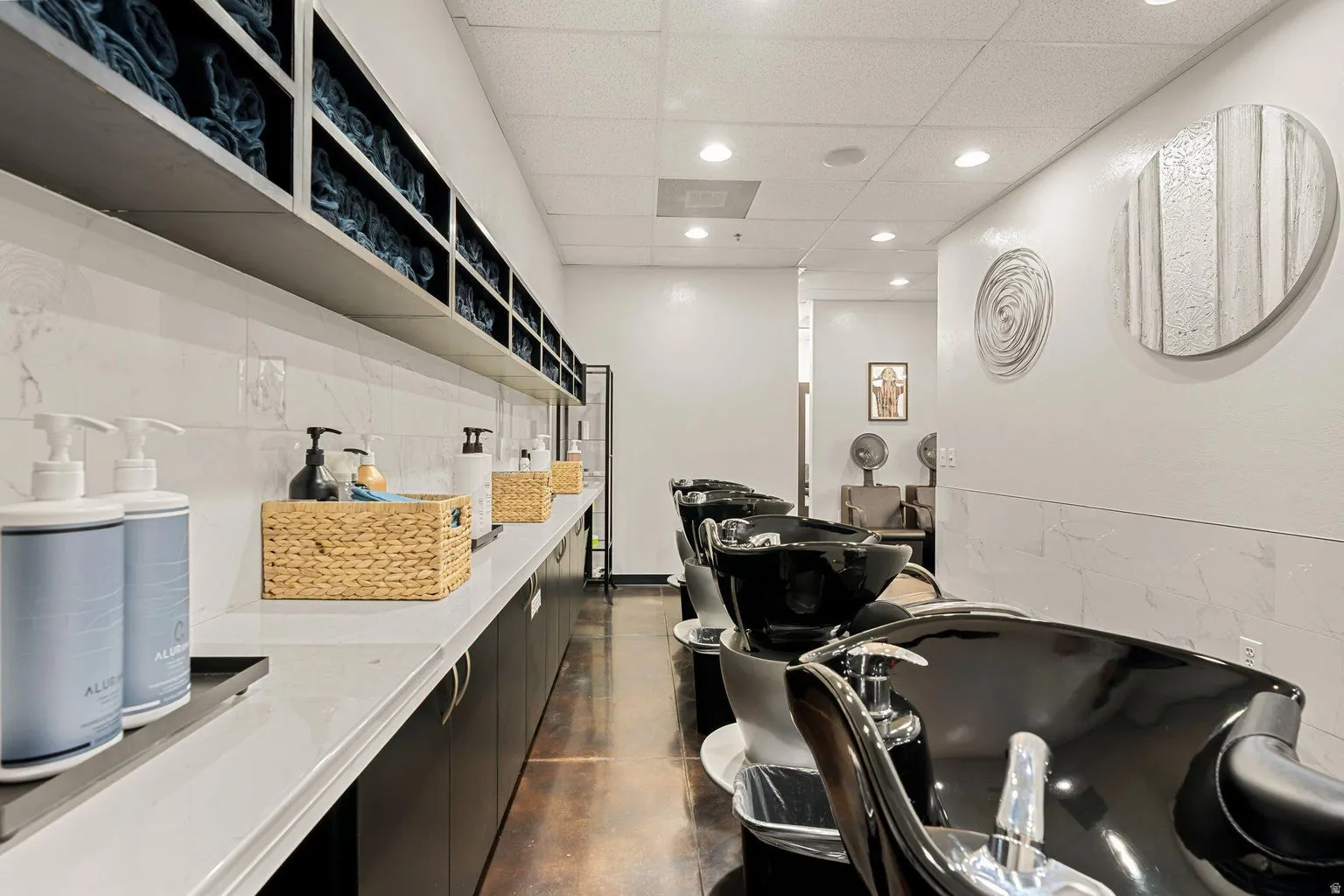 Integrated shelving unit, recessed ceiling lighting, tiled accent wall, dark wood-finish flooring, and multiple salon washing stations