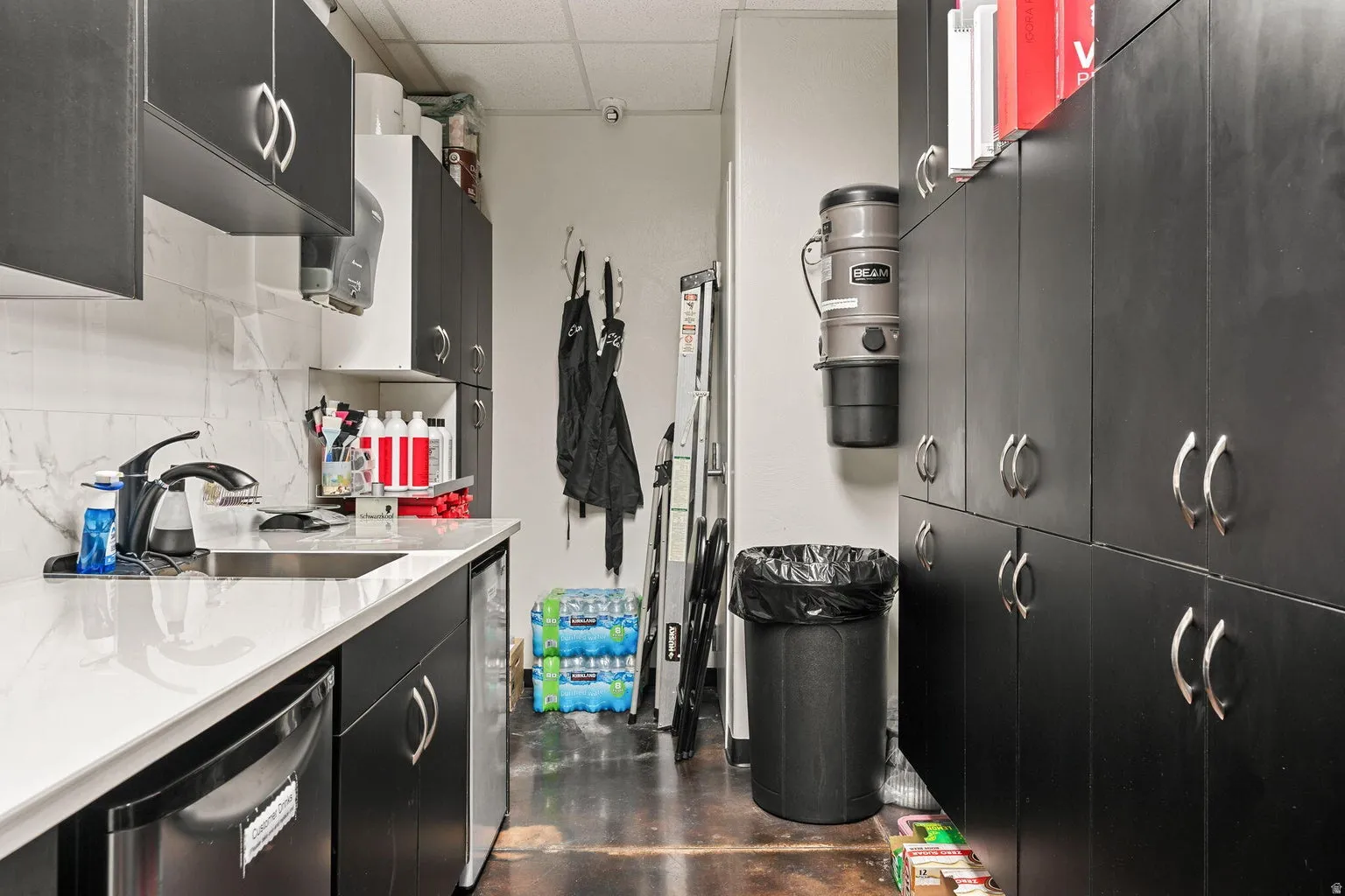 Sleek black cabinetry with chrome hardware, white solid surface countertop, stainless steel sink, and a built-in compact refrigerator
