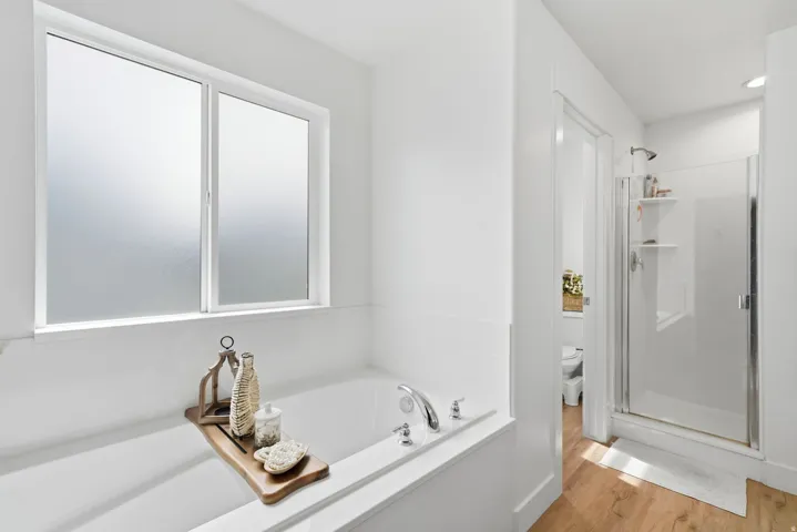 Soaking tub with deck-mounted chrome fixtures, frosted glass window, walk-in shower with glass enclosure, and wood-finish flooring