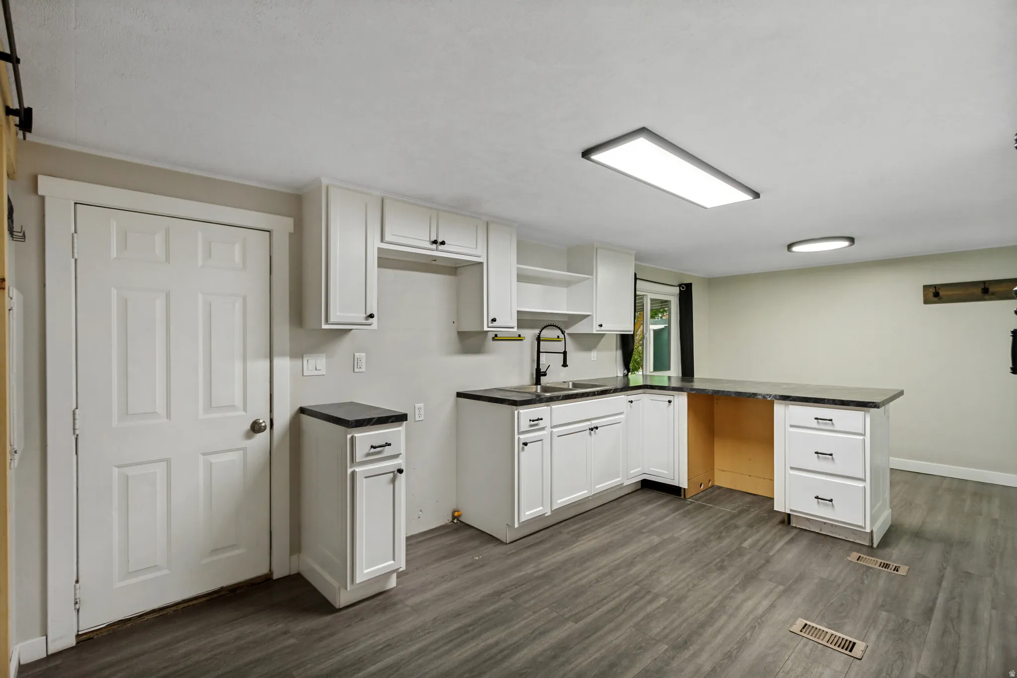 Modern kitchen featuring white cabinetry, dark countertops, a built-in sink with a black gooseneck faucet, and wood-finish flooring