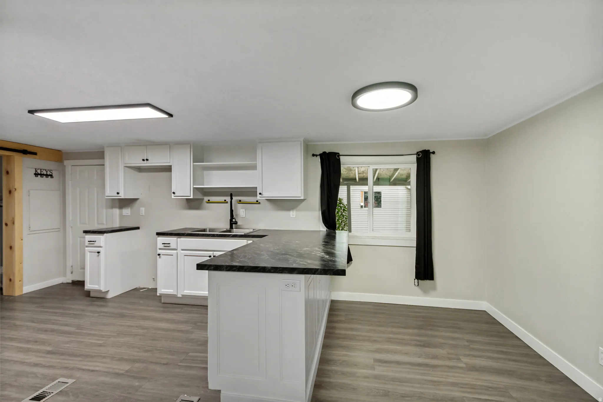 Modern kitchen featuring white cabinetry, dark countertops, wood-finish flooring, and a contemporary sink with a black faucet