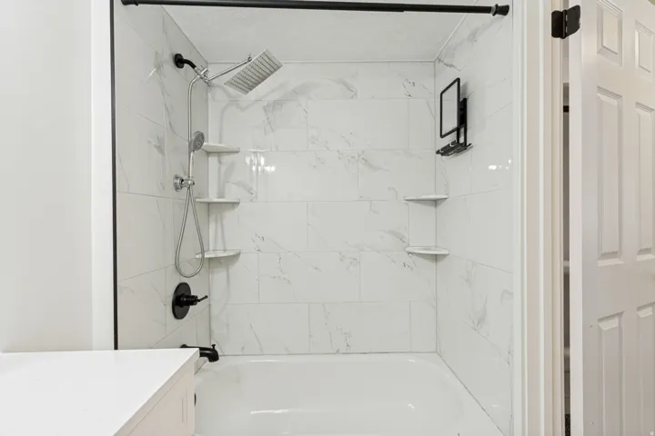 Bathtub and shower combination featuring white tiled walls, a rain showerhead, a handheld shower fixture, and built-in corner shelving