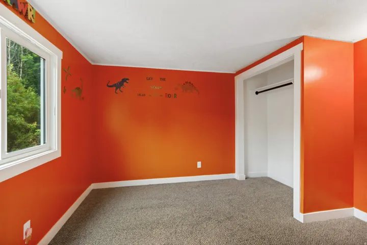 Carpeted flooring, white trim details, window with outdoor greenery, and a closet with a hanging rod