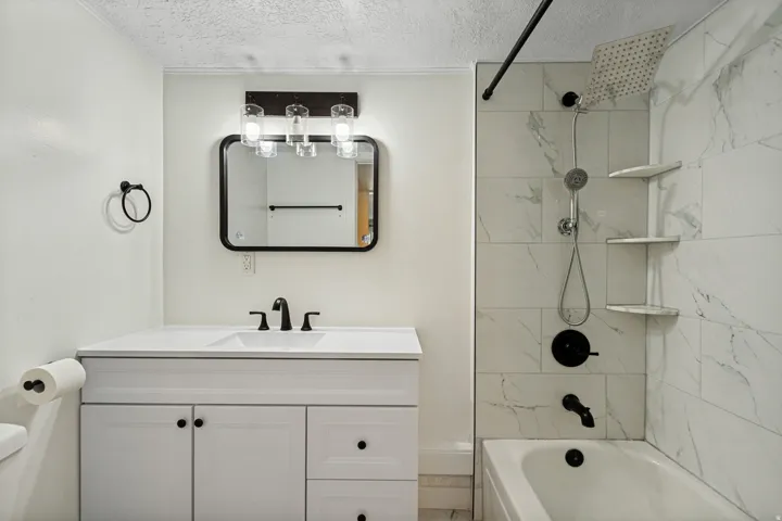 Vanity with integrated sink and matte black fixtures, framed mirror, three-light vanity bar, tiled shower surround with built-in shelving, and a rain shower head with a handheld attachment