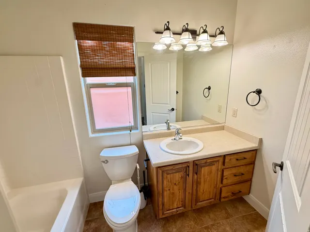 Bathroom featuring a single basin vanity with wood cabinetry, a built-in tub, a window with a woven shade, and tile flooring