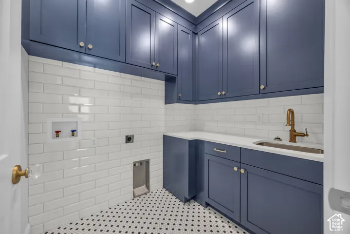Laundry room featuring full-height blue cabinetry, white subway tile backsplash, white countertops, a stainless steel sink with a bronze-toned faucet, and a patterned floor tile