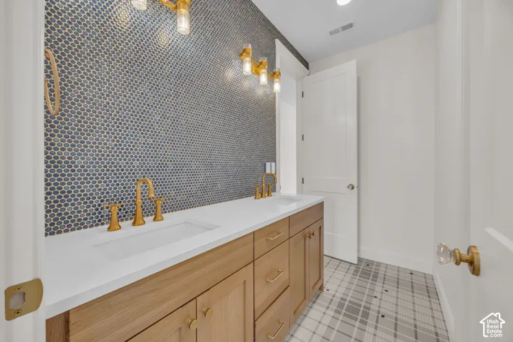 Dual vanity with integrated sinks, wood-finish cabinetry, gold-toned fixtures, mosaic tile accent wall, and patterned floor tile