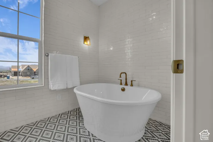 Freestanding bathtub with brass fixtures, white subway tile walls, patterned floor tiles, and a large window