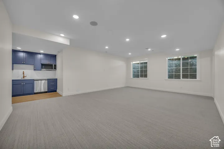 Spacious room featuring broadloom carpet, two windows, recessed lighting, and a built-in wet bar with blue cabinetry, a stainless steel dishwasher, and a microwave
