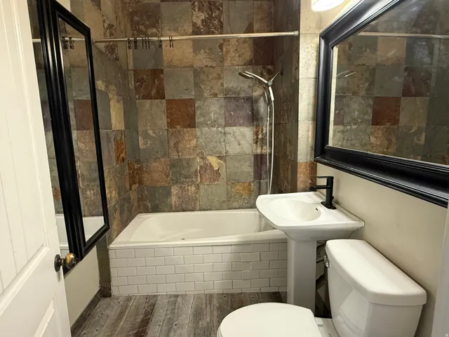 Pedestal sink with a matte black faucet, subway tile tub surround, full wall tile shower, and wood-finish flooring