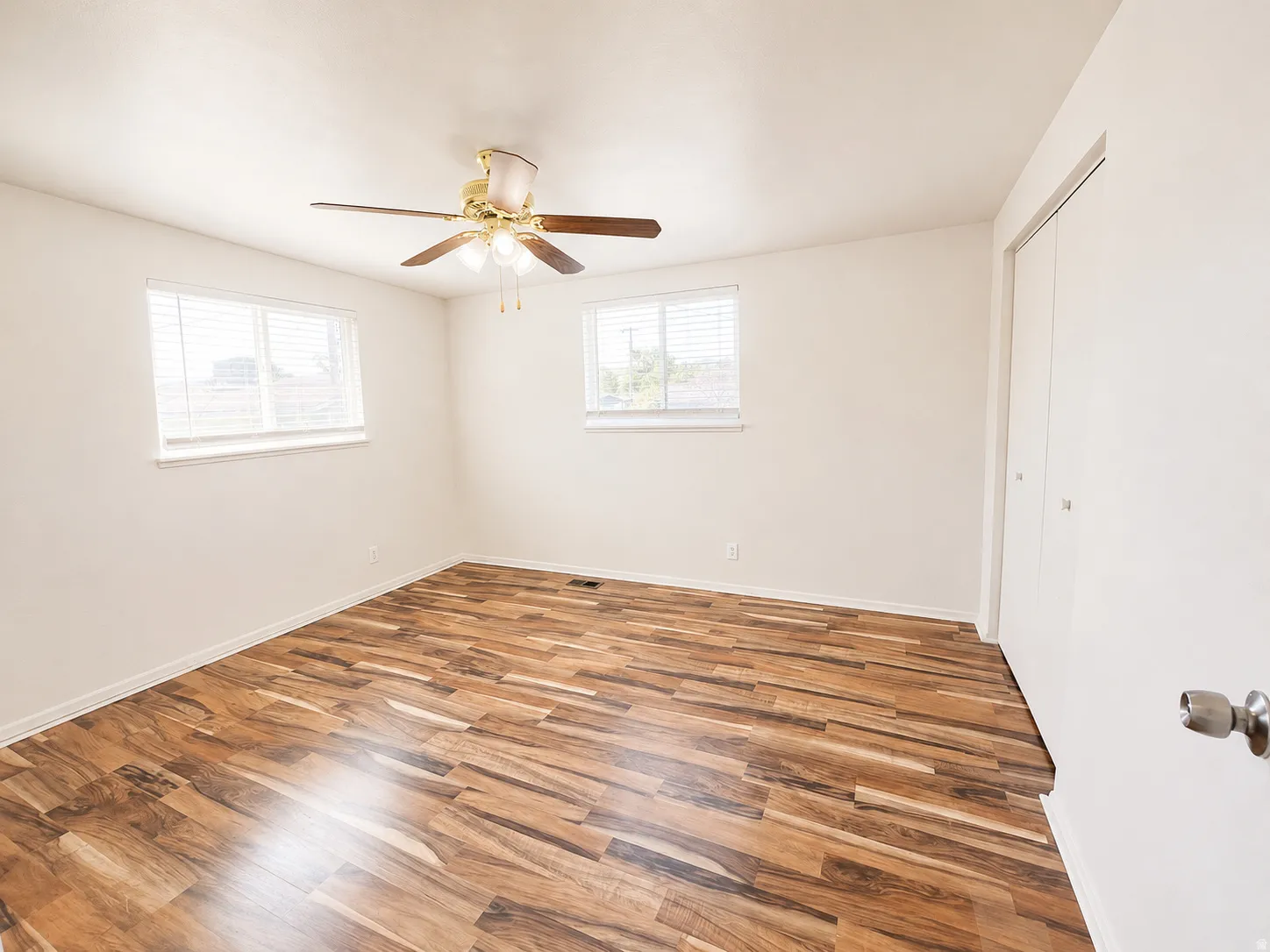 Wood-finish flooring, two windows with blinds, ceiling fan with integrated lighting, and a closet with bifold doors