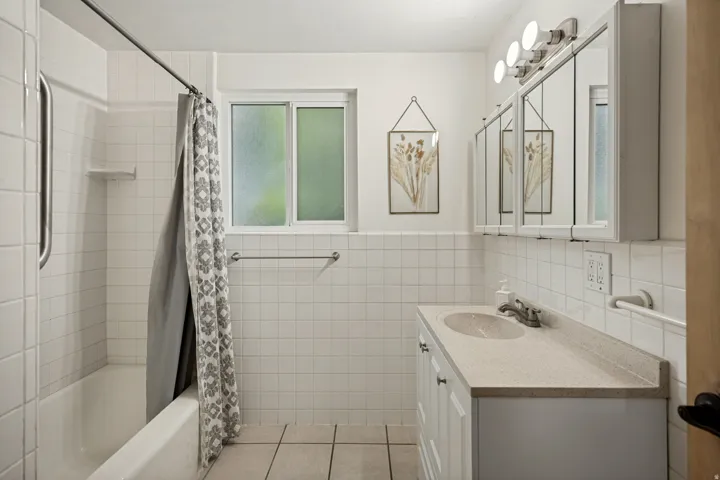 White tiled bathroom featuring a built-in bathtub, a window, a vanity with an integrated sink, and triple mirrored medicine cabinets