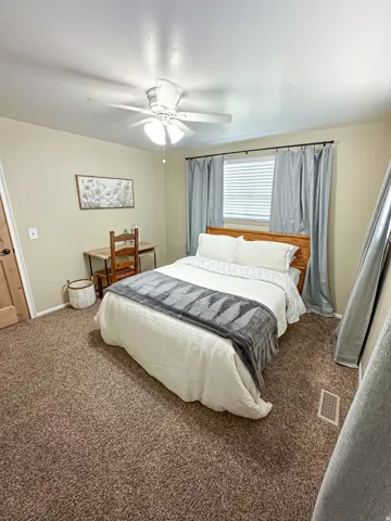 Window with horizontal blinds and drapery, overhead ceiling fan with light fixture, light brown textured carpeting, and wood-finish door