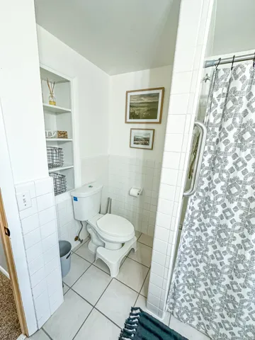 Recessed built-in shelving, white square wall tiling, white ceramic toilet, tile flooring, and a shower with a grab bar