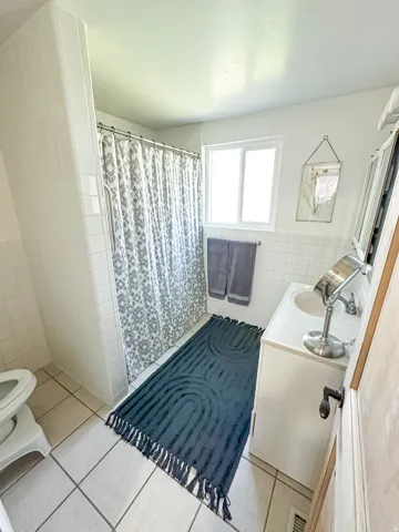 White tiled shower enclosure, white vanity with integrated sink, rectangular window, white tiled flooring, and a towel bar