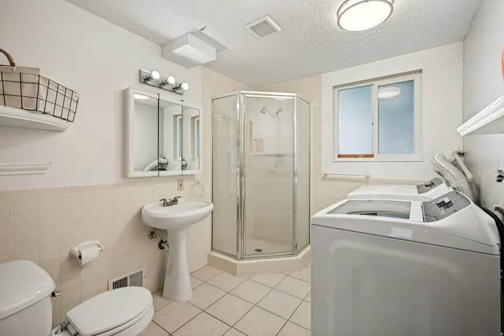 Pedestal sink, wall-mounted medicine cabinet with integrated lighting, glass-enclosed corner shower, tiled flooring, and a window with obscured glass
