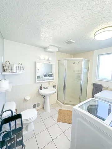 White tile flooring, pedestal sink, mirrored medicine cabinet, glass-enclosed shower, and built-in window