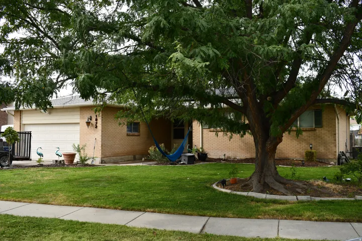 Brick exterior with an attached garage, featuring a mature shade tree, a manicured lawn, and a concrete sidewalk
