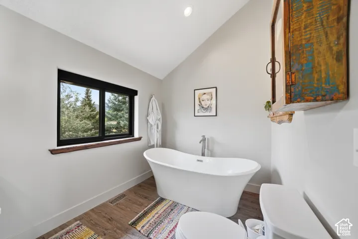 Freestanding soaking tub with floor-mounted faucet, wood-finish flooring, and vaulted ceiling