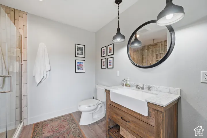 Walk-in shower with vertical tile surround, wood-finish flooring, a white farmhouse-style sink, a stone countertop, and dual pendant lighting