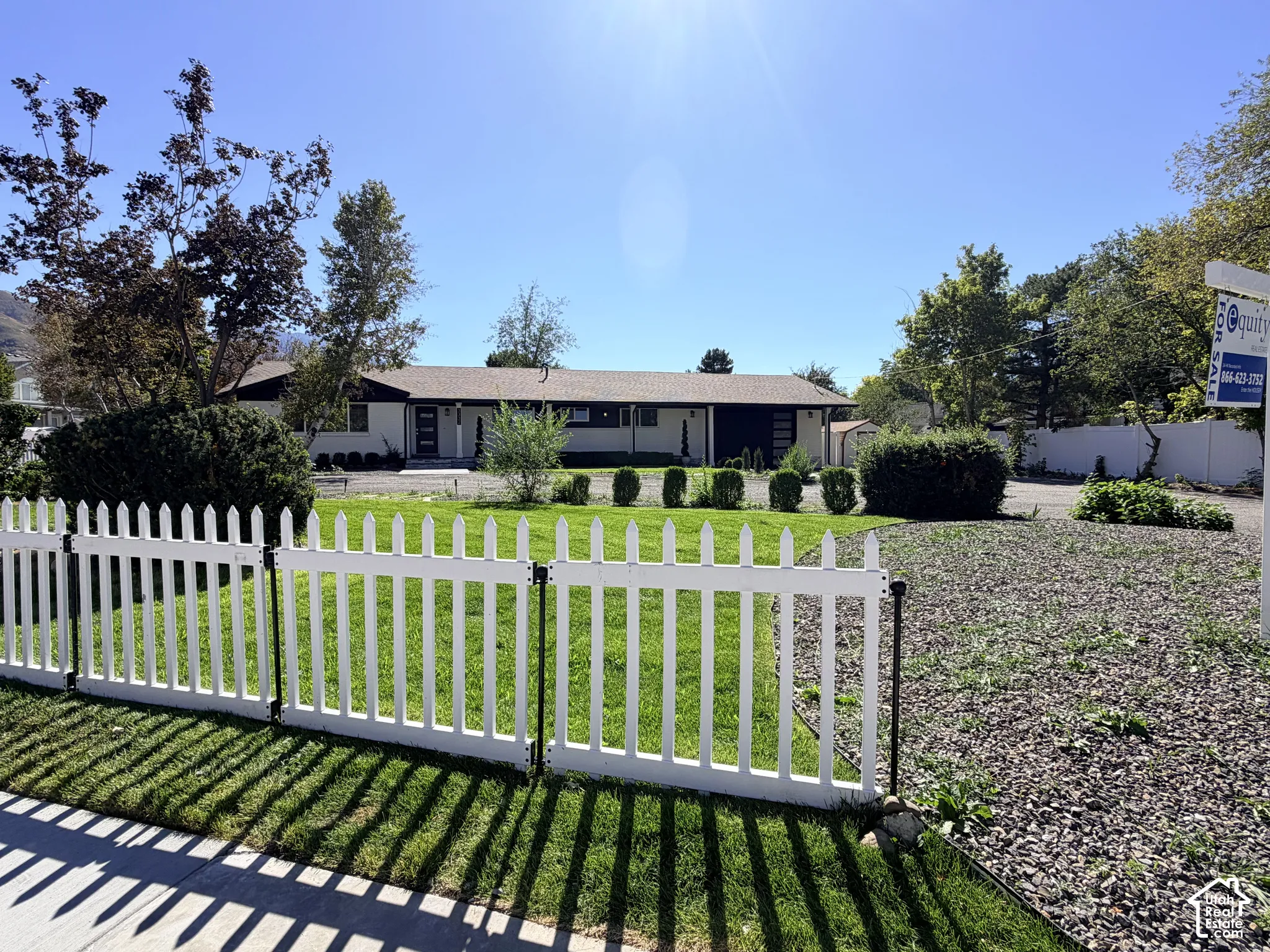 White picket fence, manicured lawn, established shrubbery, mature trees, and a gravel driveway