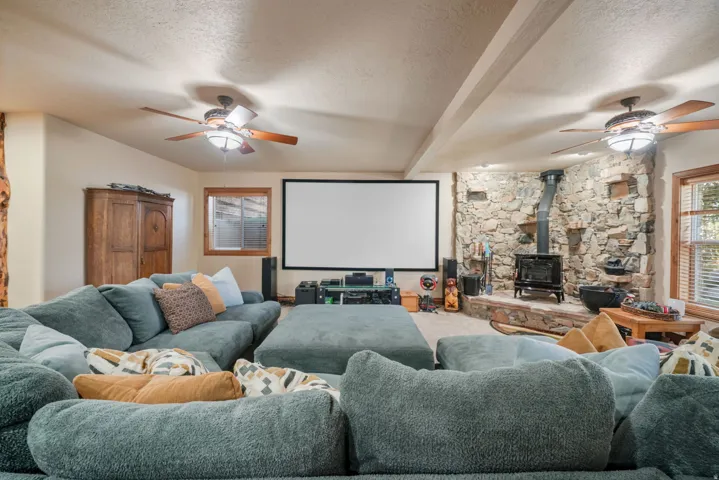 Expansive recreation room featuring multiple seating areas with built-in sectional configuration, large projection screen with mounted media console, stone fireplace with wood-burning stove, coffered ceiling with ceiling fans, and abundant natural light f
