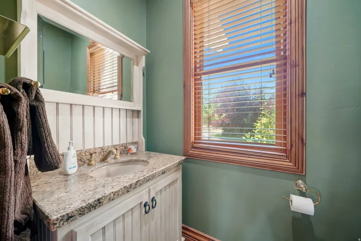Wood-framed window with blinds, vanity featuring a granite countertop, undermount sink, and brass-finish faucet